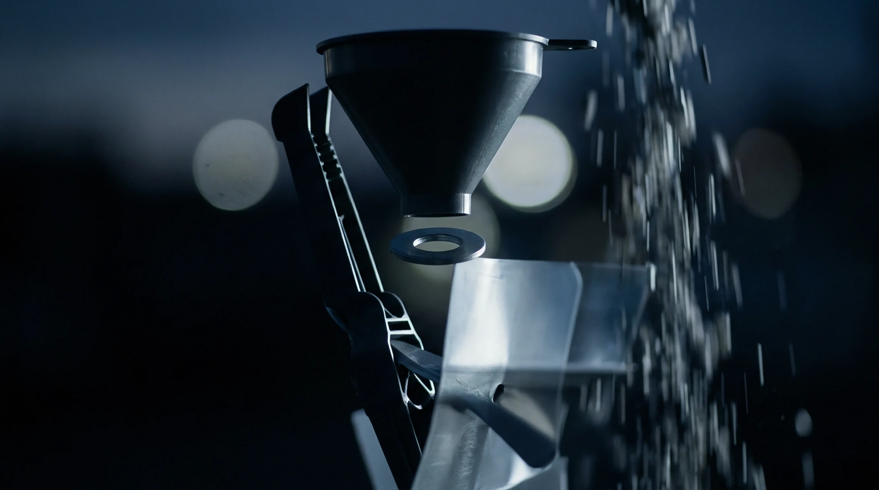 Plastic granules stream past a black funnel and a clear polymer shield mounted on a metal tooling clamp assembly.